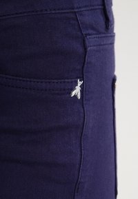 Navy blue denim with a smooth texture, featuring a side pocket and a small silver embroidered detail resembling a stylized insect.