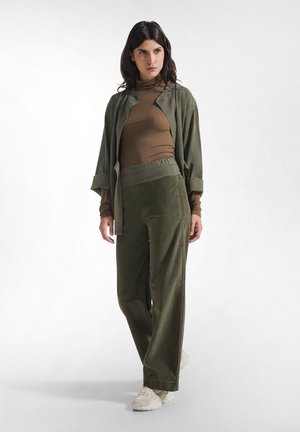 Donna con giacca verde oliva, dolcevita marrone, pantaloni in velluto a coste verde oliva e sneakers beige, in piedi davanti a uno sfondo bianco.