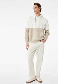 Witte en beige hoodie met een kenguruzak, gecombineerd met lichte beige broek en crèmekleurige sneakers, staand tegen een neutrale achtergrond.
