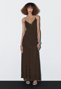Woman with curly hair wearing a long brown sleeveless dress and silver sandals, standing against a white background with silver bracelets.