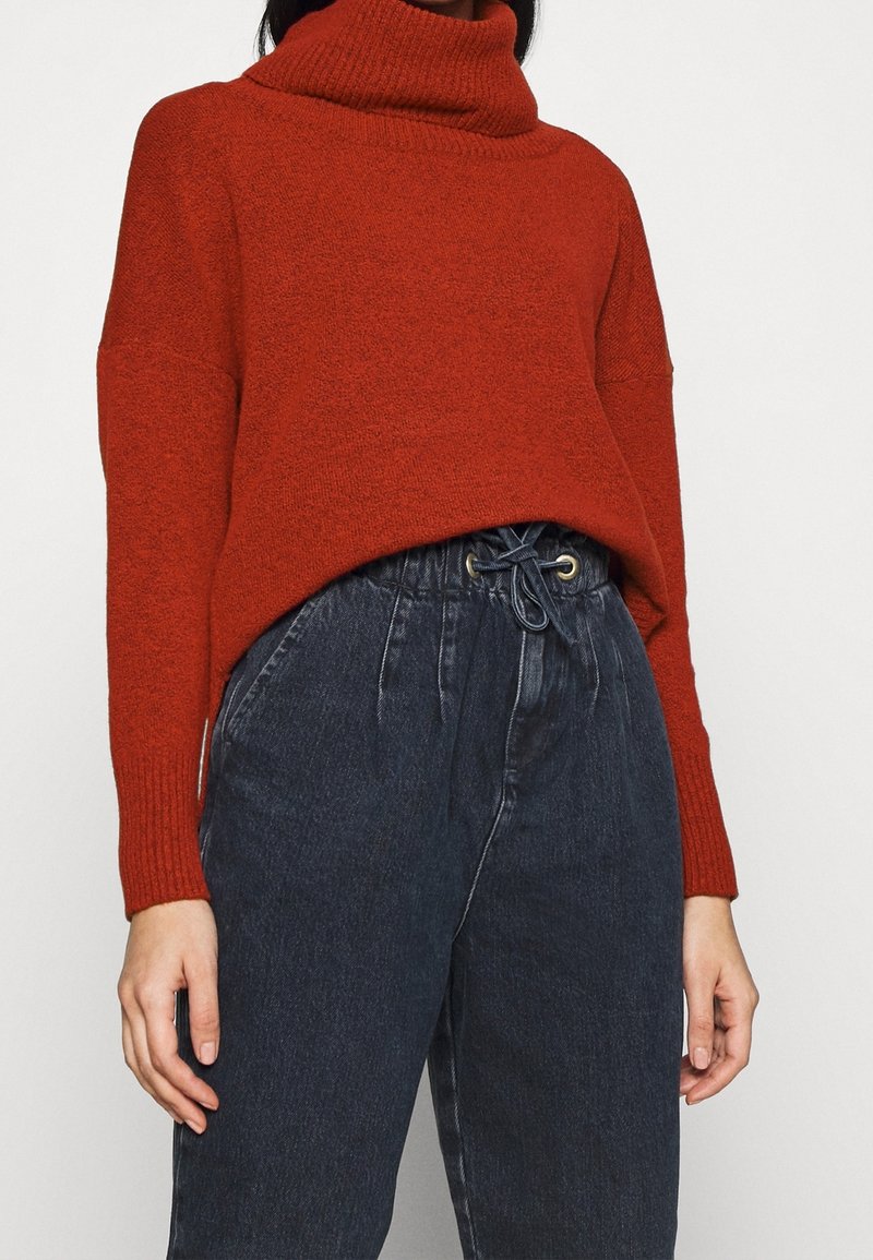 Maglione rosso a maglia con colletto alto e vestibilità ampia, abbinato a pantaloni in denim scuri a vita alta con vita elastica.