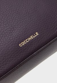 Purple textured leather pouch with embossed "Coccinelle" logo in gold. Smooth edges and minimalistic design.