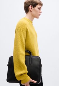 Cartable en cuir noir avec une poignée épaisse, de forme rectangulaire et une surface texturée, associé à un pull jaune en tricot.