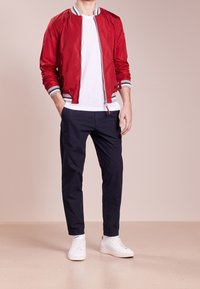 Uomo che indossa una giacca bomber rossa, una t-shirt bianca, pantaloni blu marino e sneakers bianche, in piedi con le mani in tasca su uno sfondo beige.