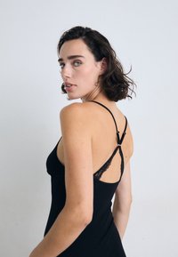 Zwart mouwloos jurkje met kantaccenten op de schouderbanden, aansluitend ontwerp en een gladde textuur. Vanop een zijaanzicht gedragen, waardoor de details aan de achterkant worden benadrukt.