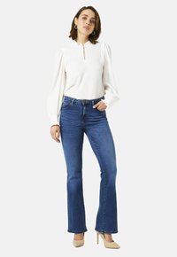 Vrouw in een witte blouse met lange mouwen, blauwe hoge taille bootcut jeans, beige hoge hakken en gouden hoepeloorbellen, staand met haar hand in haar zak.