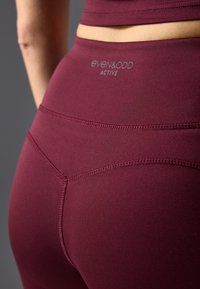 Burgundijske aktivne tajice s glatkom teksturom, s logotipom na pojasu i detaljima šavova koji naglašavaju prilagođeni oblik.