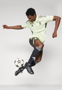 Polo shirt in lichtgroen met zwarte accenten, logo op de borst, gecombineerd met zwarte shorts en sokken. Speler in beweging met een voetbal.