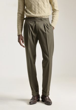 Pantalones verde oliva con pliegues, ajuste entallado y un detalle de cintura alta. Combinados con un suéter de punto de cable color crema y zapatos marrones.