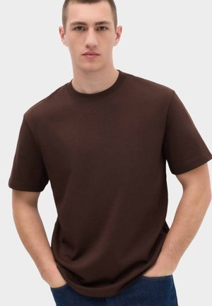 Młody mężczyzna w gładkim ciemnobrązowym t-shircie z krótkim rękawem i niebieskich dżinsach, z rękami w kieszeniach na jasnym tle.