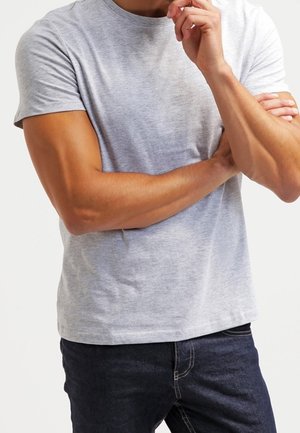 Mann in hellgrauem kurzärmligen T-Shirt und dunkler Jeans, die Arme verschränkt, eine Hand berührt das Kinn, vor schlichtem Hintergrund.