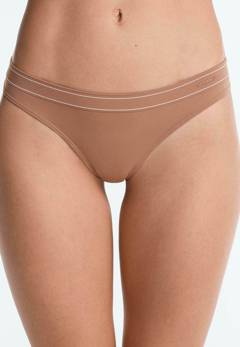 Gros plan sur le bas du torse d’une femme portant un sous-vêtement beige sans couture de style hipster avec une fine ceinture à double rayure.