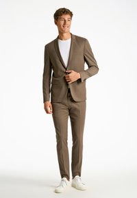 Completo marrone a due pezzi con vestibilità su misura, blazer a un bottone, pantaloni dritti e una maglietta bianca, abbinato a sneakers bianche.