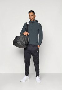 Meeste spordiriietus: tumehall kapuutsiga jakk, mustad dressipüksid valge Nike logoga, valged spordijalatsid, käes must spordikott.