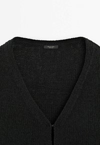 Cardigan noir en maille avec un col en V, texture visible et une petite étiquette rectangulaire de la marque au niveau de l'encolure.
