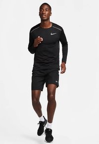 Mężczyzna w czarnej koszulce Nike z długim rękawem, czarnych szortach i czarnych butach biegowych biega na tle jednolitego białego tła.