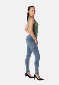 Vaqueros ajustados de color azul con cintura alta, hechos de mezclilla, con dos bolsillos traseros y una pequeña etiqueta con logo. Combinados con una blusa verde sin mangas y tacones negros.