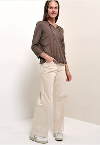 Femme portant un sweat à capuche marron à manches longues, un pantalon large beige et des baskets blanches, debout avec une main dans la poche.