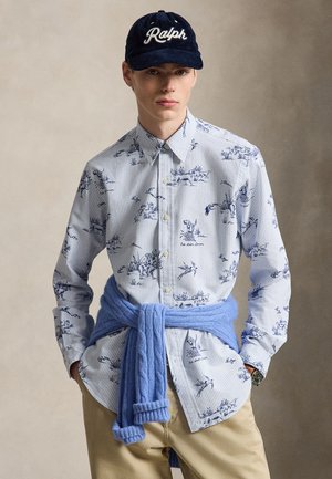 Jeune homme portant une chemise à motifs boutonnée, un pantalon beige, une casquette bleu marine avec "Ralph," et un pull bleu noué autour de la taille.