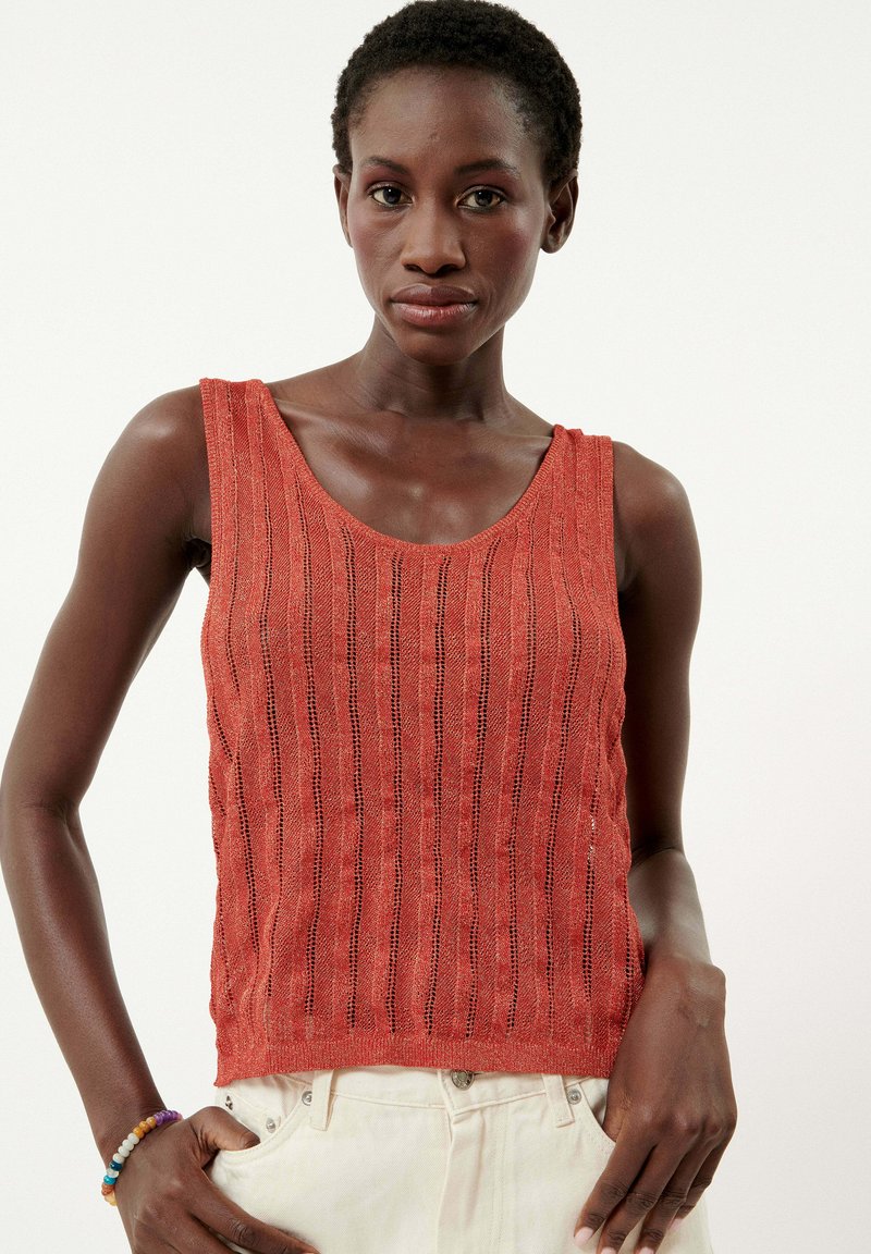 Débardeur en tricot rouge avec un motif de rayures verticales, encolure dégagée et texture en maille ouverte. Porté avec un pantalon de couleur claire, présentant une poche visible.