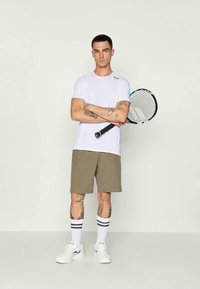 T-shirt atlético branco, calções khaki, ténis brancos e meias às riscas. Segurando uma raquete de ténis, de pé com os braços cruzados.