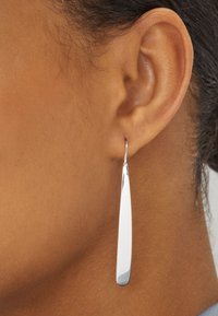 Boucles d'oreilles pendantes en argent avec une forme allongée et élégante et une surface polie, suspendues à un simple crochet. Peau texturée et détails de l'oreille visibles.