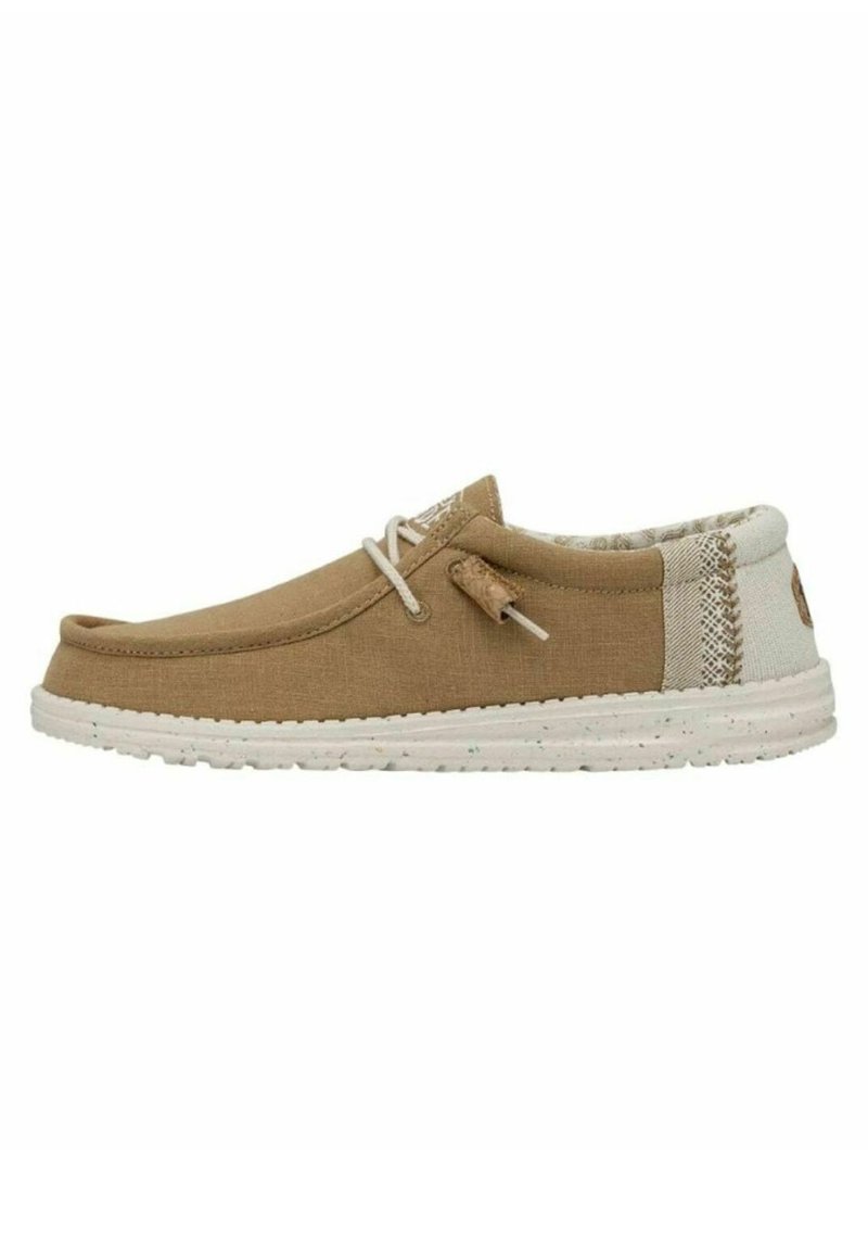 Zapato slip-on de lona marrón con suela de goma blanca, que presenta un panel trasero texturizado y un detalle decorativo de cordones en la parte delantera.