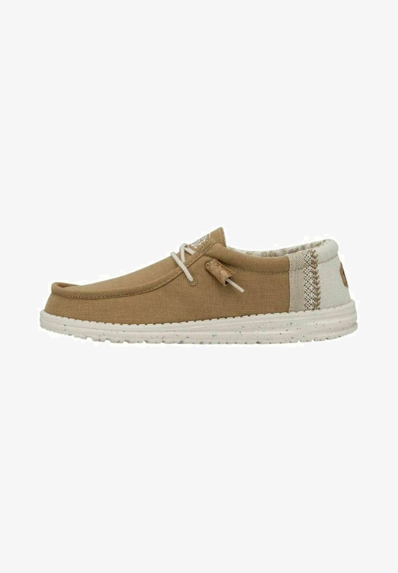 Zapato slip-on de lona marrón con suela de goma blanca, que presenta un panel trasero texturizado y un detalle decorativo de cordones en la parte delantera.