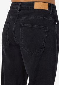 Crne traperice visokog struka s džepovima na stražnjoj strani. Kožni brand patch na pojasu. Glatka tekstura i ravni kroj nogavica.