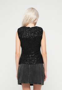 Mulher com cabelo loiro longo, vestindo uma parte de cima sem mangas preta com lantejoulas e uma saia de jeans pregueada escura, vista de costas contra um fundo branco.