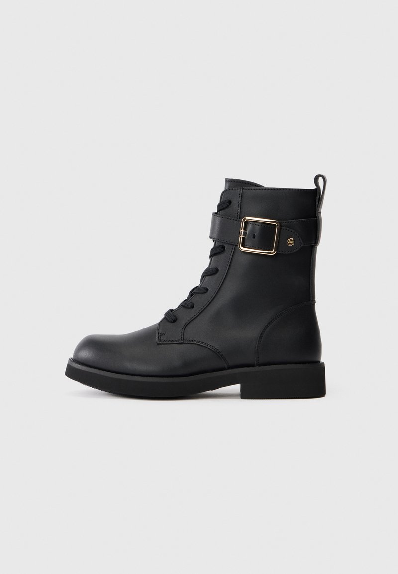 Stivaletto in pelle nera con lacci frontali, ampia fascia con fibbia dorata e linguetta per il tallone sul retro, su uno sfondo chiaro e semplice.