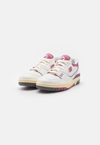 Baskets New Balance blanches avec des accents violets. Les caractéristiques incluent une tige en cuir lisse, des panneaux texturés et une semelle en caoutchouc beige.