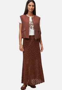 Gilet sans manches marron avec fermeture éclair sur un t-shirt blanc à motif graphique, associé à une longue jupe en dentelle marron et des sabots à clous.