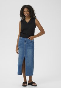 Top sans manches noir associé à une longue jupe en denim bleu avec une fente frontale, portée avec des sandales à glisser noires.