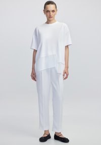 Blusa blanca de capas con mangas cortas, combinada con pantalones blancos. Zapatos planos negros con acentos decorativos. Textura de tela suave.