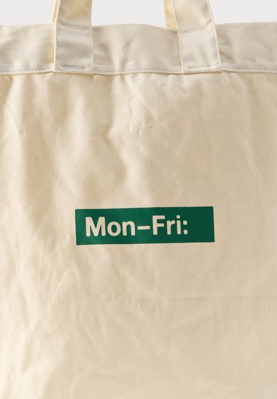 Geantă tote din pânză bej cu mânere rezistente și o casetă verde cu textul „Lun–Vin:” în centrul frontal.