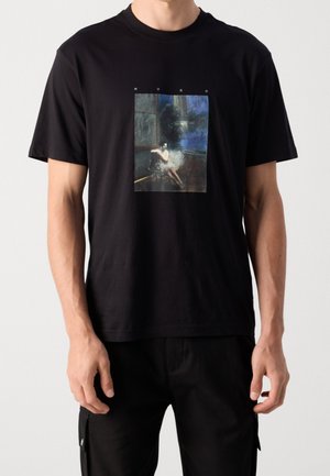 Mężczyzna ubrany w czarny T-shirt z kolorowym nadrukiem malarskim ukazującym siedzącą postać, nad obrazem znajduje się napis "HUGO".