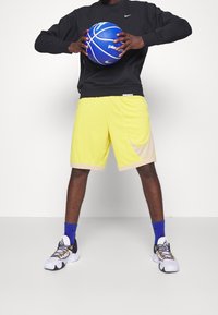 Sudadera negra, pantalones cortos amarillos con acentos beiges, balón de baloncesto azul y zapatillas negras/blancas con detalles grises y calcetines azules.
