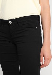Schwarze Denim-Jeans mit schmalem Schnitt, ausgestattet mit einer Vordertasche, silbernem Knopfverschluss und gestickten Details. Das Material ist glatt mit leichtem Stretch.