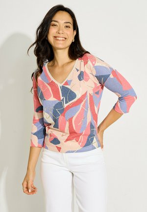 Mujer sonriendo, vistiendo una camiseta de cuello en V con un patrón abstracto y colorido, mangas tres cuartos y pantalones blancos, posando con una mano en la cadera.