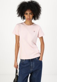 Tricou din bumbac roz deschis, cu mâneci scurte, guler rotund și un mic logo triunghiular pe pieptul stâng, purtat împreună cu o pereche de pantaloni din denim albastri.