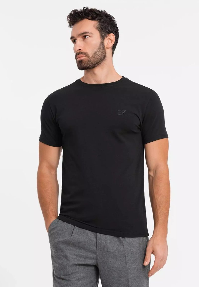 Sort kortærmet t-shirt lavet af blødt stof, med et diskret logo på brystet og en klassisk pasform. Båret med grå bukser.