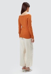 Oranges Strickpullover mit langen Ärmeln, tiefem Ausschnitt im Rücken, kombiniert mit creamfarbenen, weiten Hosen und grauen, hohen Schuhen.