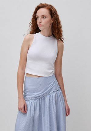 Ruda kobieta z kręconymi włosami nosi biały bezrękawnik typu crop top oraz niebiesko-białą spódnicę w paski z wysokim stanem na jednolitym tle.