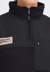 Schwarzer Fleece-Pullover mit hohem Kragen, der einen halben Reißverschluss auf der Vorderseite, eine Reißverschlusstasche auf der Brust und ein Etikett mit der Aufschrift „DESIGNED WITH DEDICATION“ aufweist.