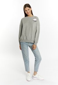 Felpa grigia con maniche lunghe e polsini a coste, caratterizzata da un design di nuvole ricamate sul lato sinistro del petto. Abbinata a jeans azzurri e sneakers bianche.