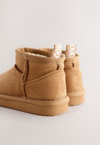 Stivaletti alla caviglia in pelle scamosciata color tabacco con morbida fodera, dotati di passanti intrecciati e cuciture testurizzate. La suola è piatta e leggermente imbottita.