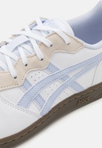 Adidași din piele albă cu accente albastre deschise, detalii din microfibră și perforații. Prezintă șireturi albe și talpă din cauciuc maro. Logo-ul ASICS este vizibil.