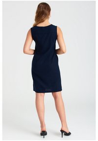 Tyrkysové šaty bez rukávů s hladkým povrchem, přiléhavou siluetou a detaily švu vzadu. Nosené s černými vysokými podpatky.