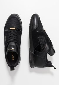 Paire de baskets noires Cruyff avec panneaux en mesh et cuir, lacets, et marquage doré sur la languette et la semelle intérieure, vues de dessus et de côté.